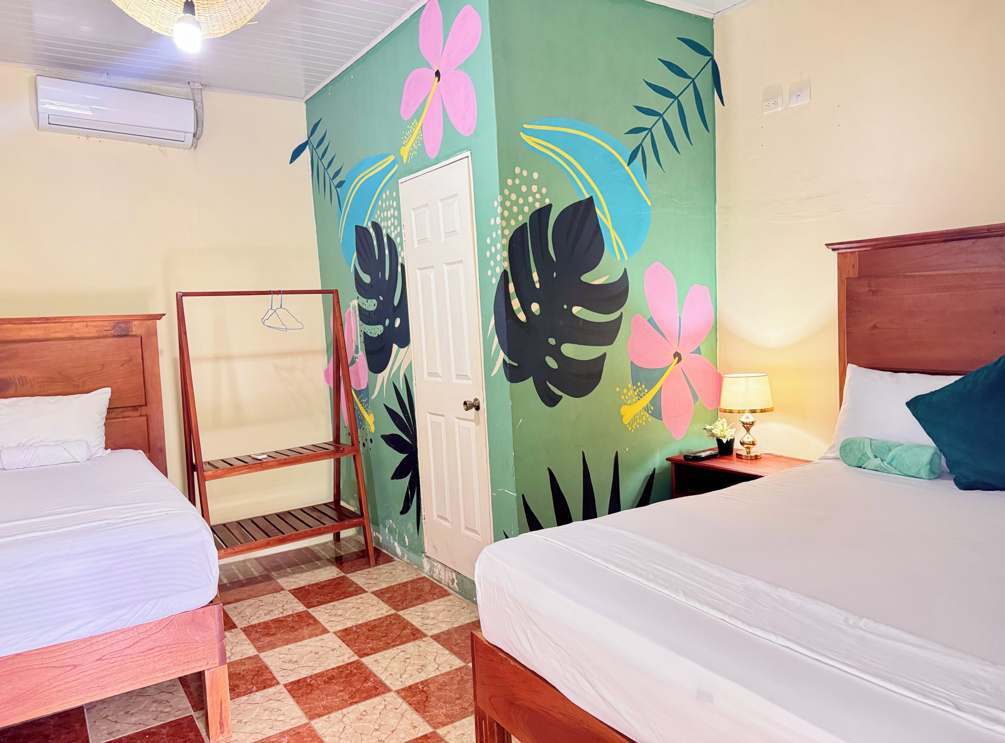 Une chambre d’hôtel avec deux lits, une tête de lit en bois, un sol à carreaux et une porte ornée d’une fresque florale.