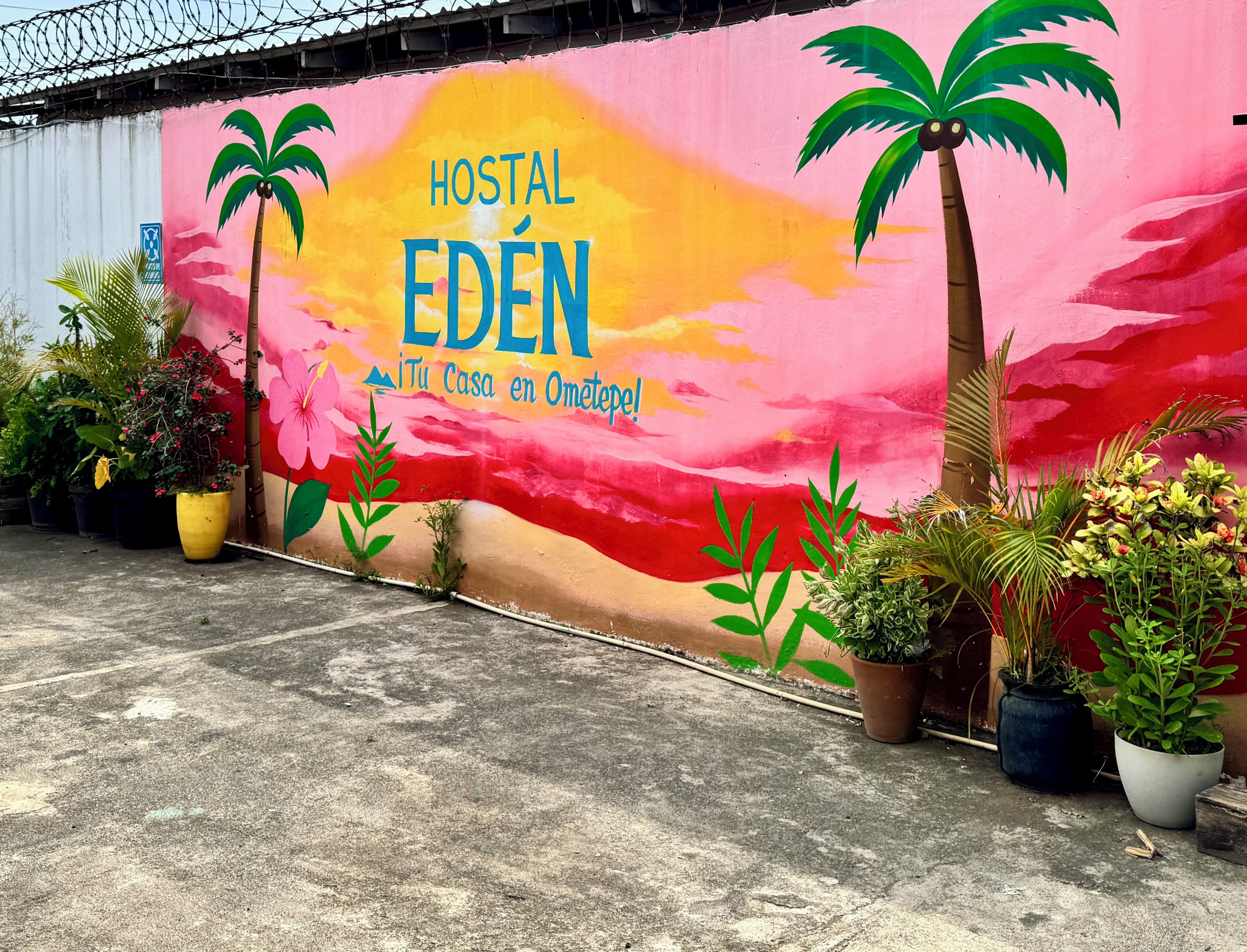 A colorful mural with the text 'HOSTAL EDEN' and 'Tu Casa en Ometepe' on a building wall, surrounded by potted plants.
