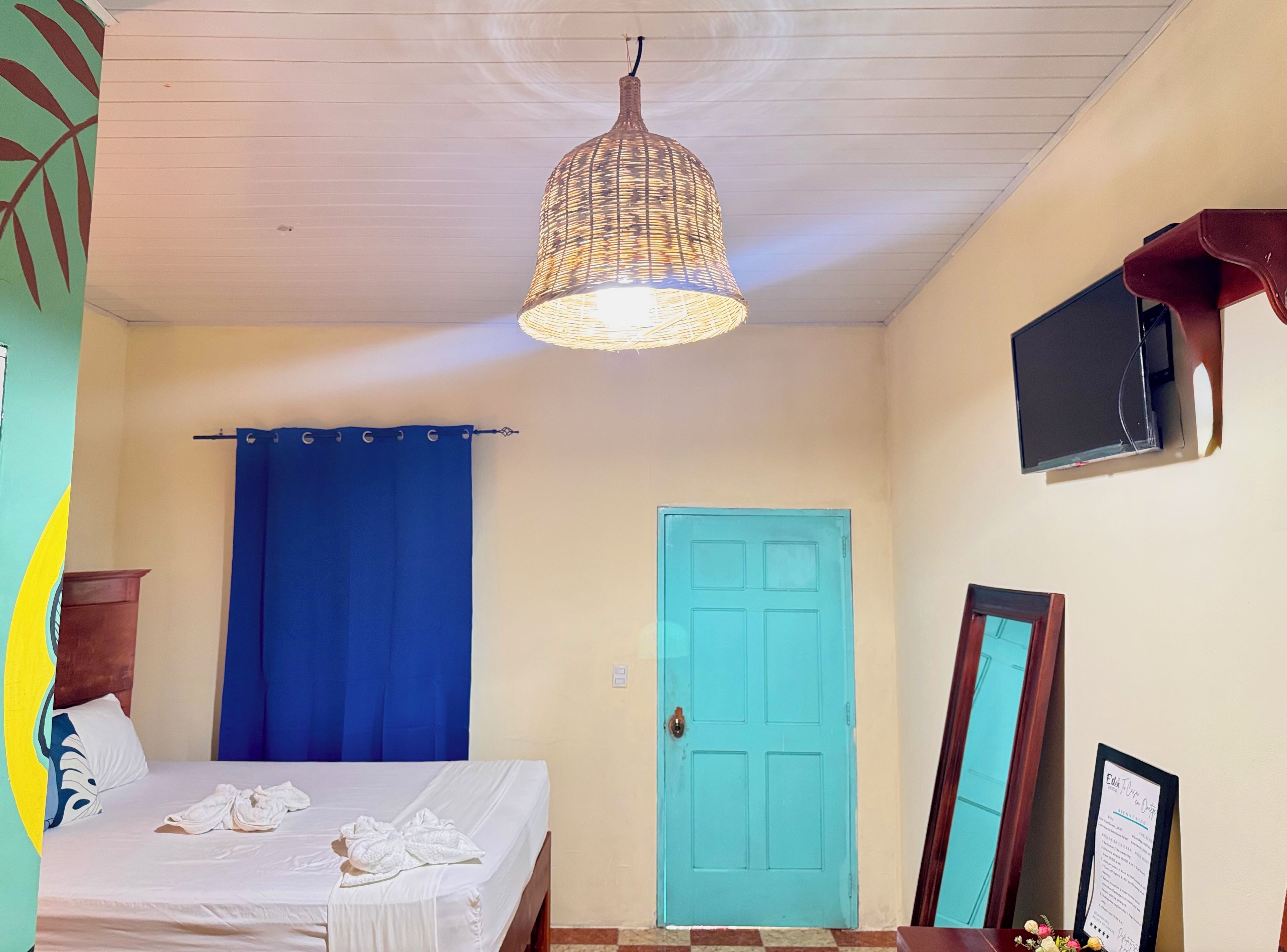 Een kamer met een bed, een blauw gordijn, een blauwgroene deur, een hanglamp, een aan de muur bevestigde televisie en een inlijsting certificaat op een voet.