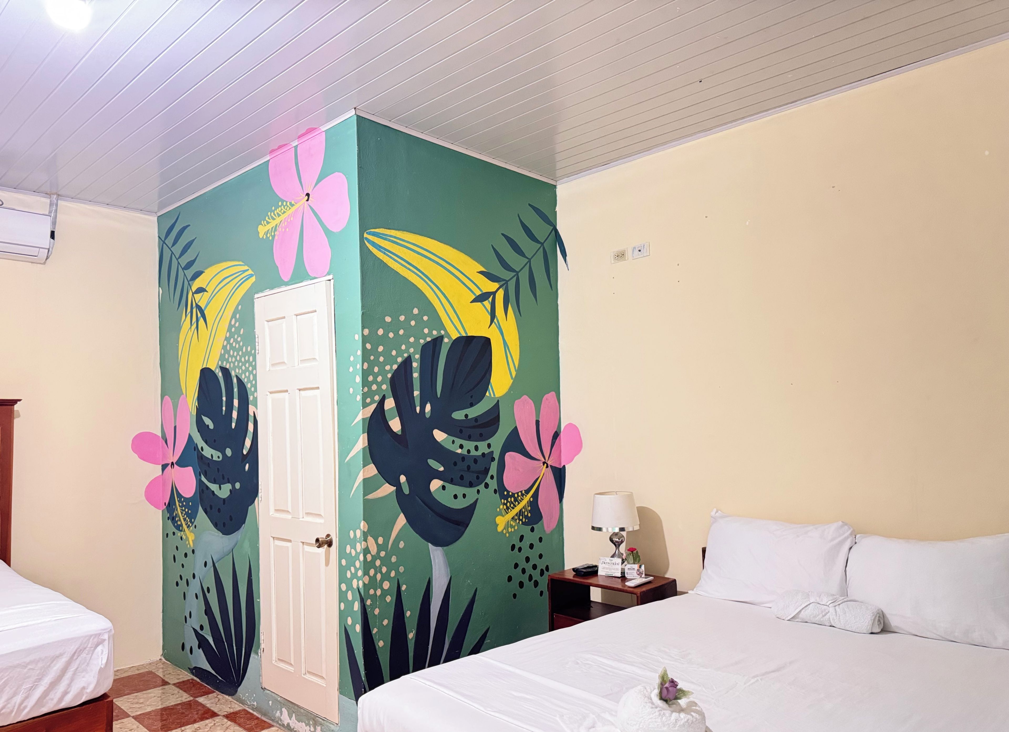 A hotel room with a bed, a nightstand, a lamp, and a wall with a tropical mural.