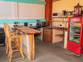 Una cocina con barra de madera, sillas, microondas, licuadora y refrigerador lleno de bebidas.