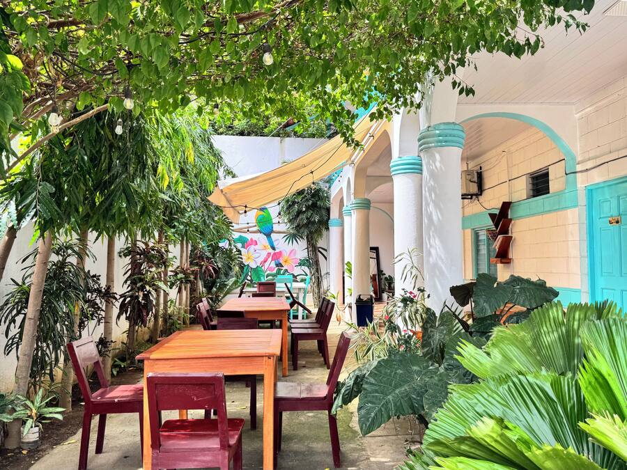 An outdoor dining area with tables and chairs, surrounded by lush greenery and a mural of a bird and flowers.