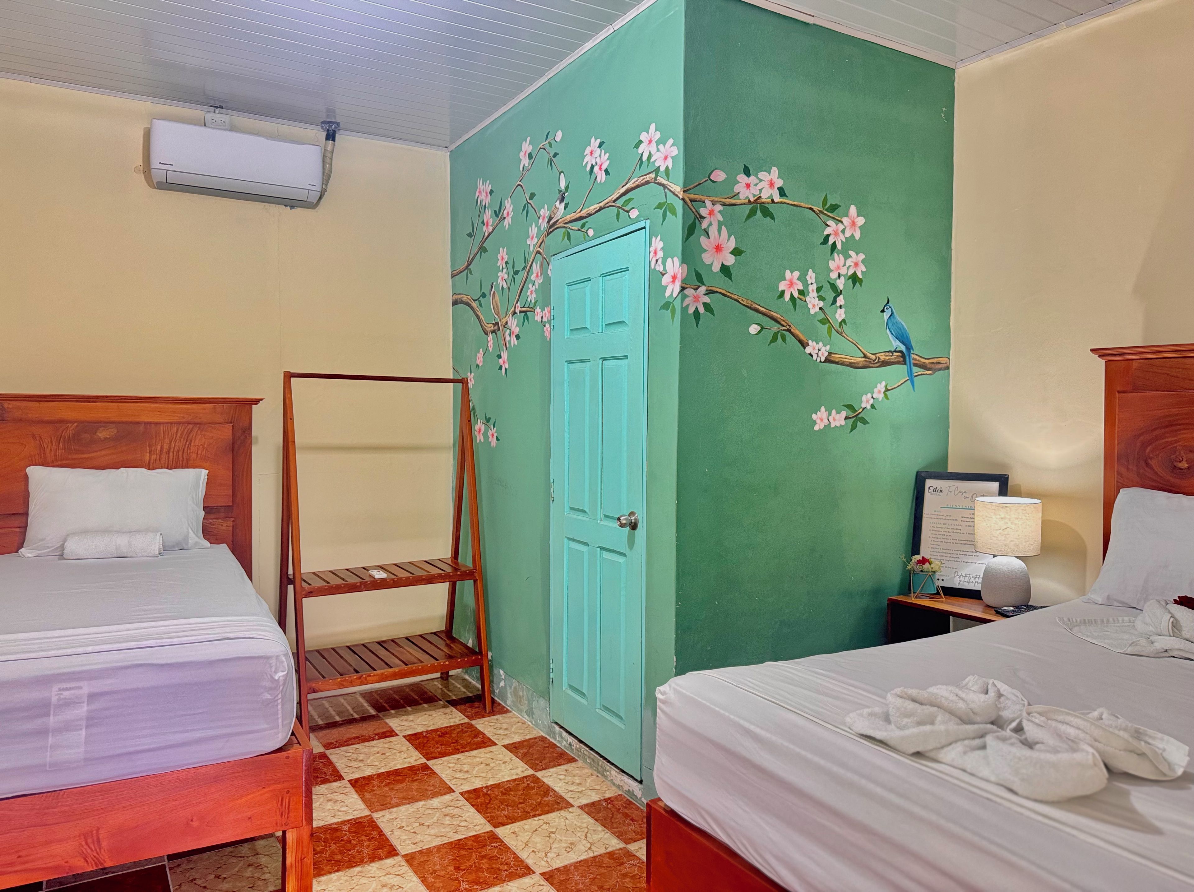 Ein Hotelzimmer mit zwei Betten, einem Schachbrettmuster-Boden, einer türkisblauen Tür und einer Wand mit floralen Bildern.