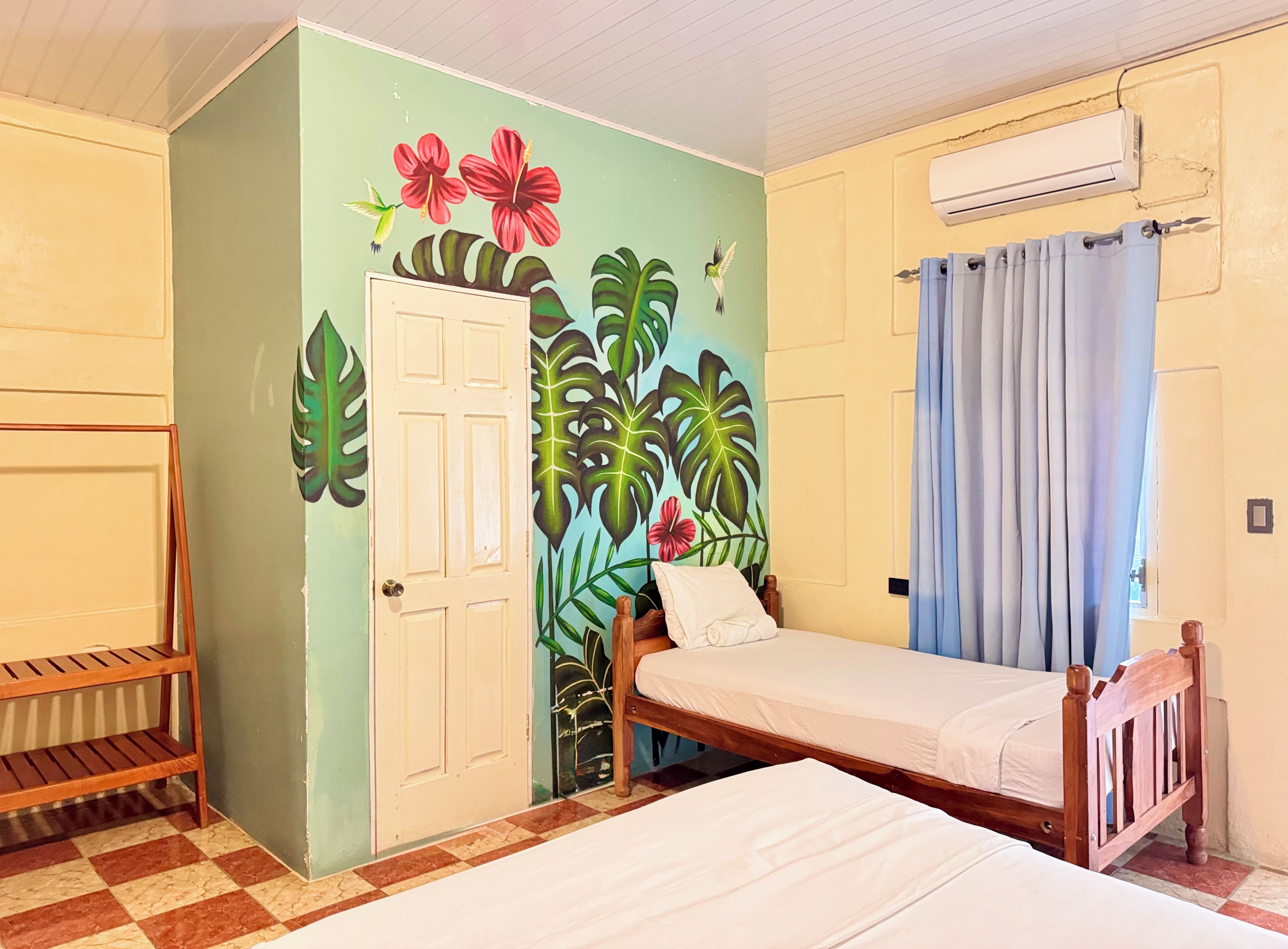 A room with a bed, a wooden shelf, a door, and a wall with a tropical mural.