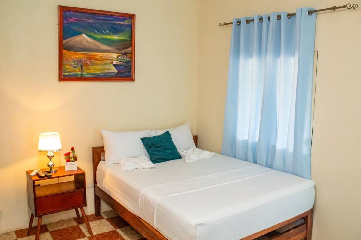 Een hotelkamer met een bed, een nachtkastje met lamp, een schilderij aan de muur en een raam met gordijnen.