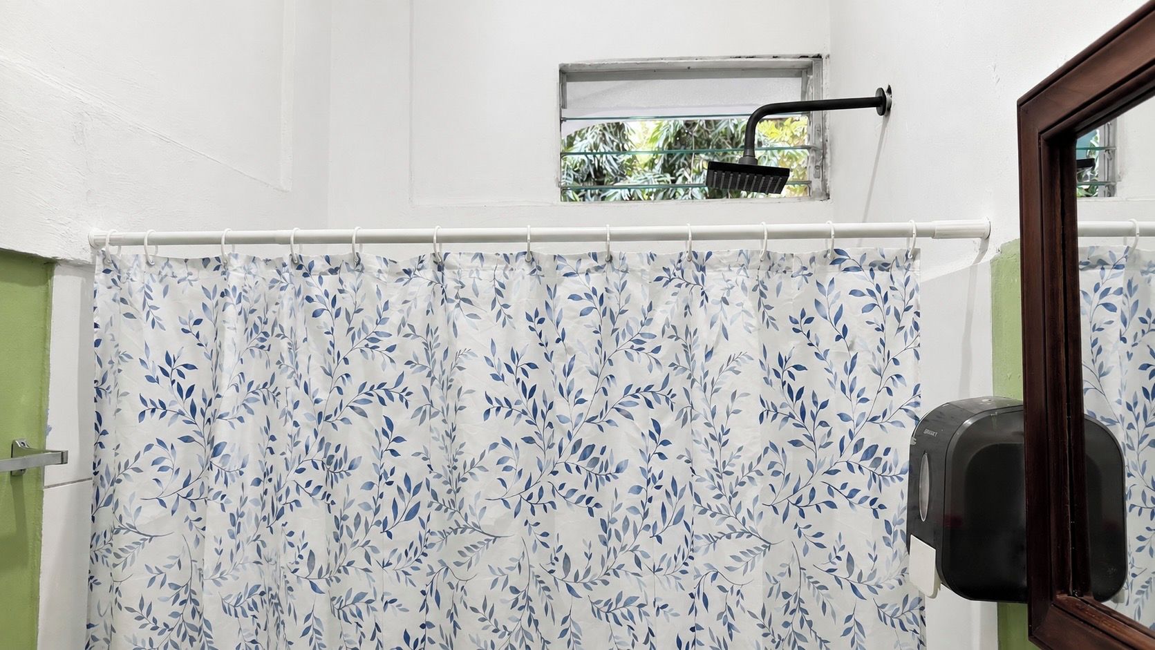 A bathroom with a shower curtain featuring a leaf pattern, a window with a view of greenery, and a hand dryer mounted on the wall.