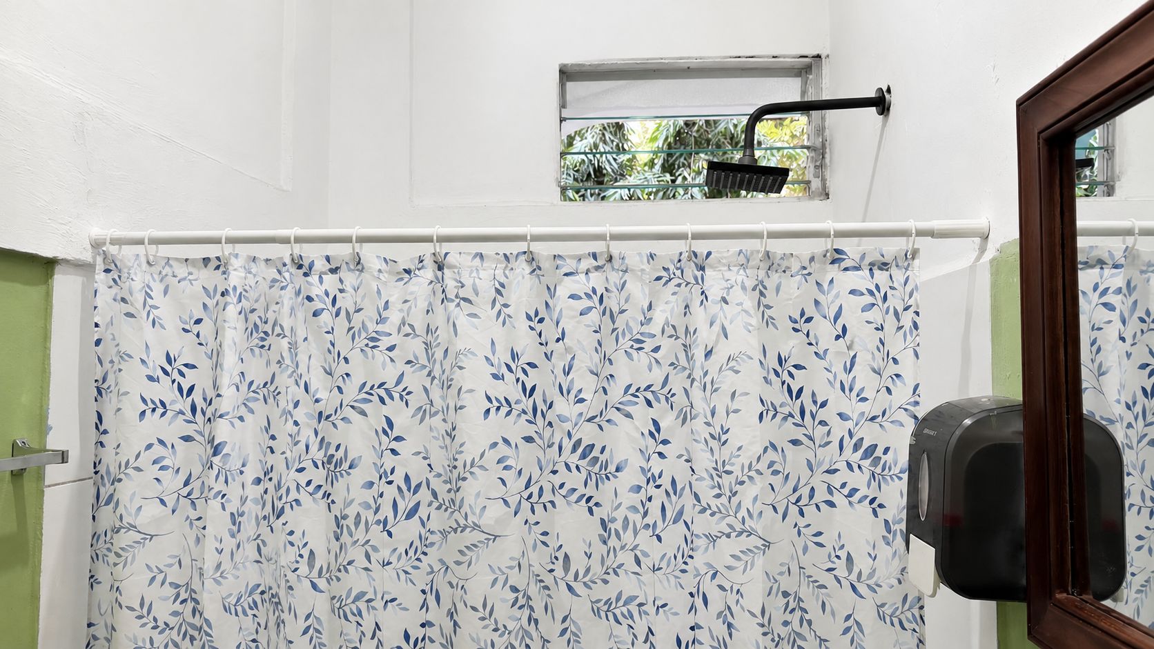 A bathroom with a shower curtain featuring a leaf pattern, a window with a view of greenery, and a hand dryer mounted on the wall.