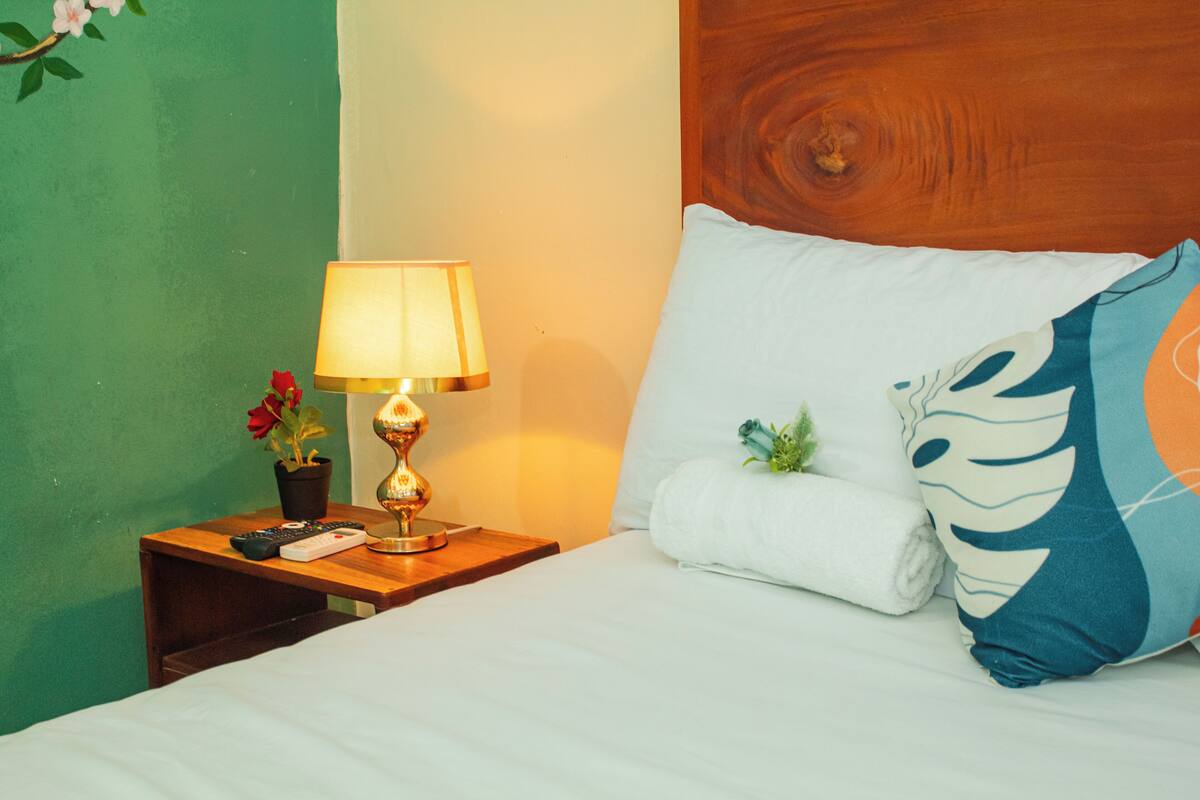 Een net opgemaakt bed met een wit kussen en een decoratief kussen, een bedlampje en een kleine potplant op een houten nachtkastje.