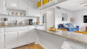 A modern kitchen with white cabinets, a granite countertop, and a coffee machine. There is a blue sofa and a TV in the living area.