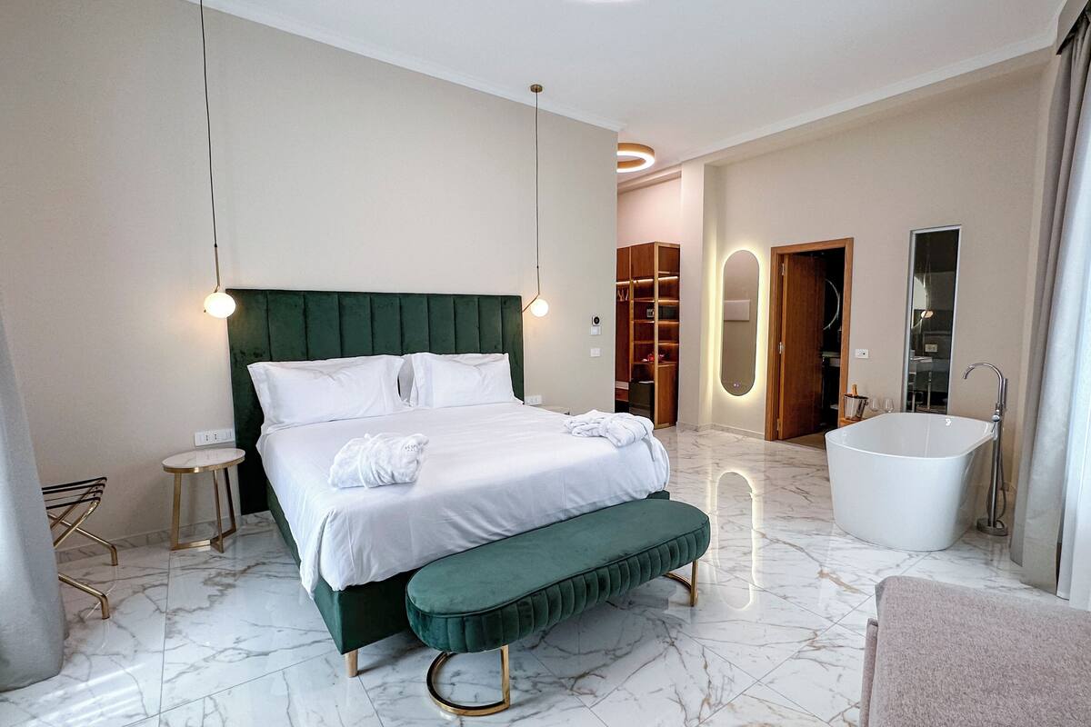 A modern hotel room with a large bed, a bench, a bathtub, and a walk-in closet.