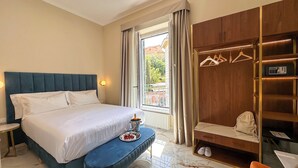 A hotel room with a large bed, a bedside table, a small bench with a tray of fruit and glasses, a wardrobe with a mirror and shelves, and a window with a view of buildings. - Vespucci Palace (Rome)