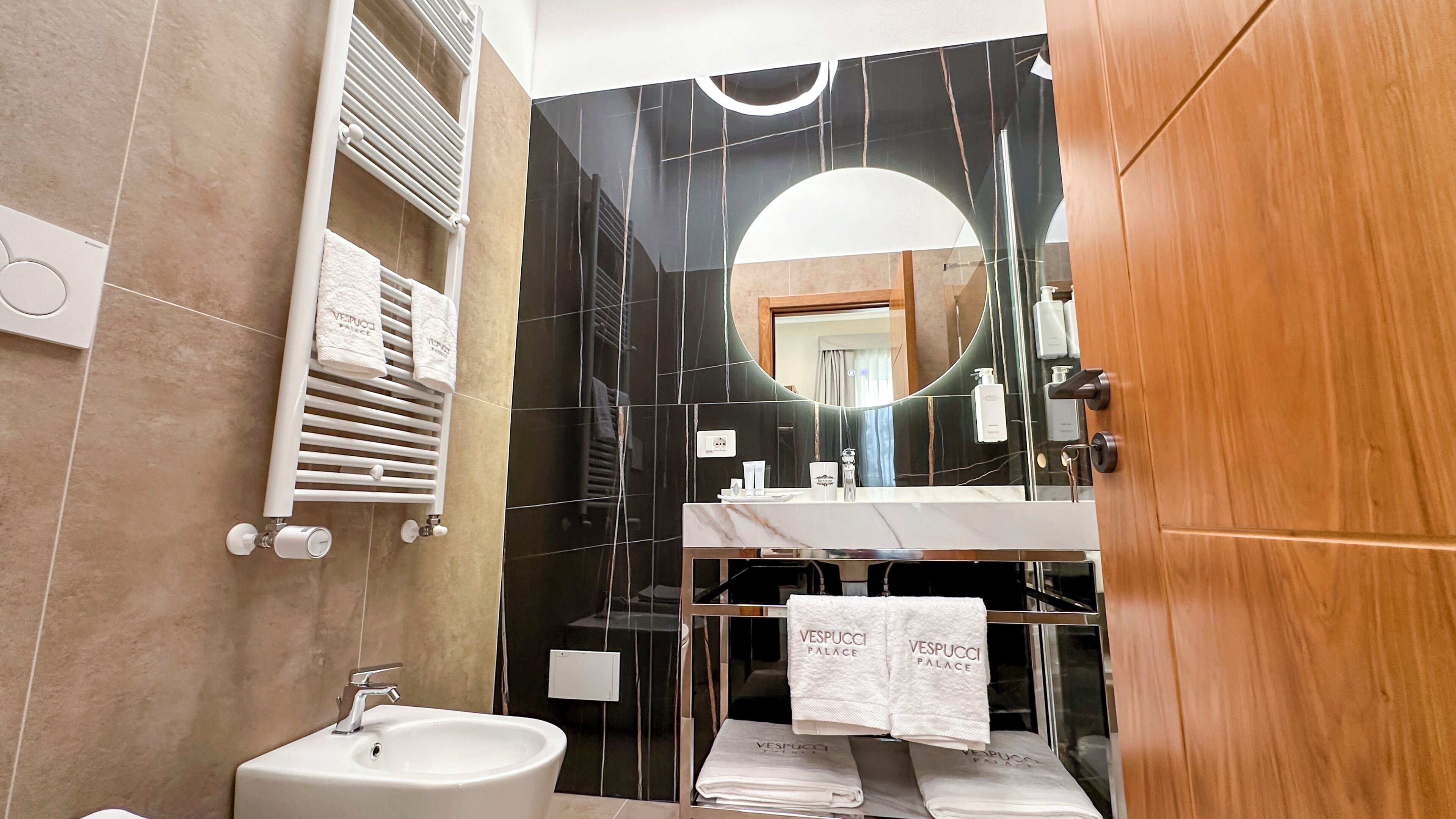 Un baño moderno con un lavabo blanco, baldosas de mármol negro, un espejo grande y una puerta de madera.