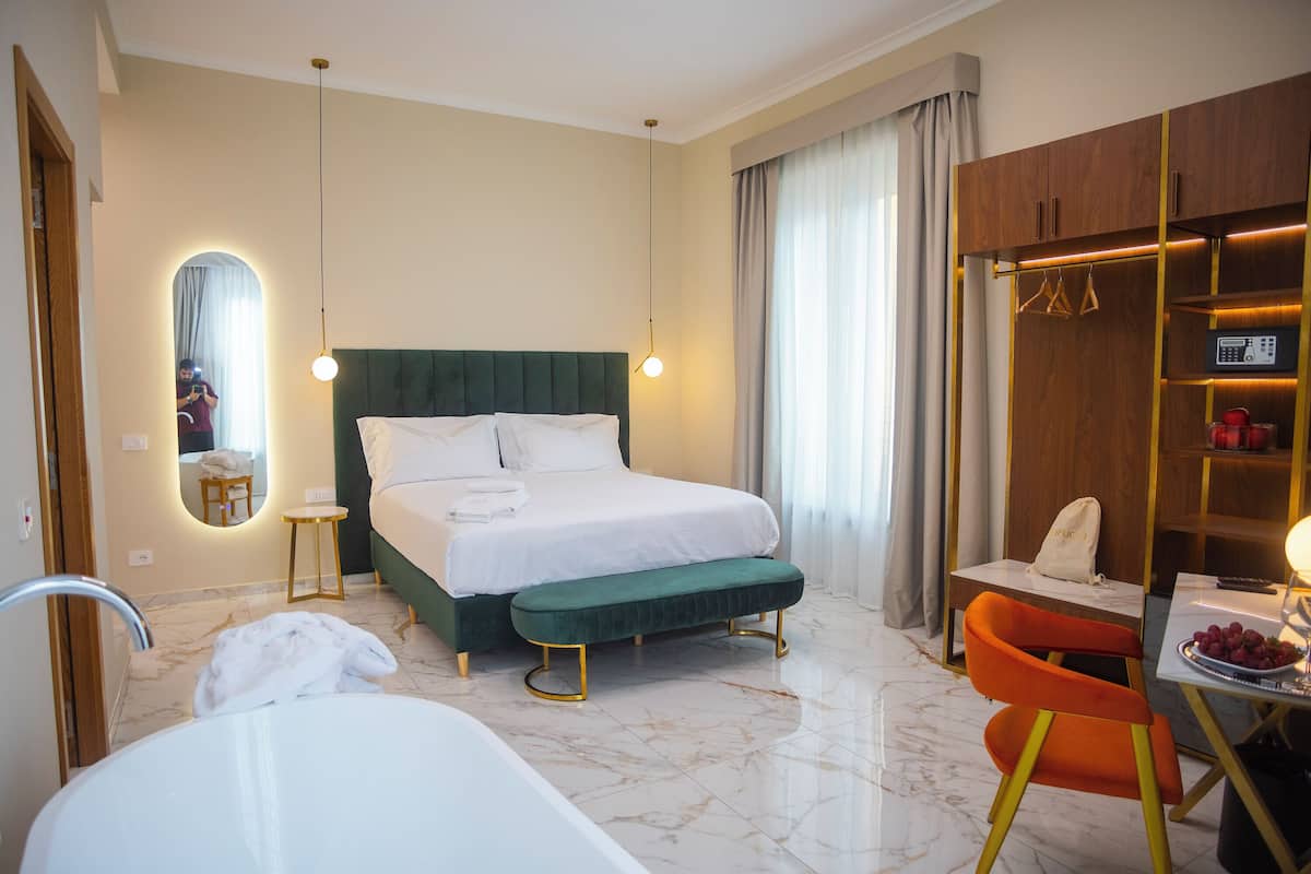 A hotel room with a bed, a bench, a chair, a table with fruit, a mirror, and a wardrobe.