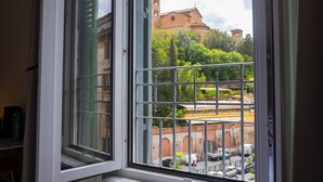 A view from a window with metal bars, overlooking a street with parked cars and a building with a clock tower. - Vespucci Palace (Rome)