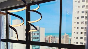 A view through a window with a metal frame and a spiral design, overlooking a cityscape with multiple buildings and a clear sky. - Co-House (João Pessoa)