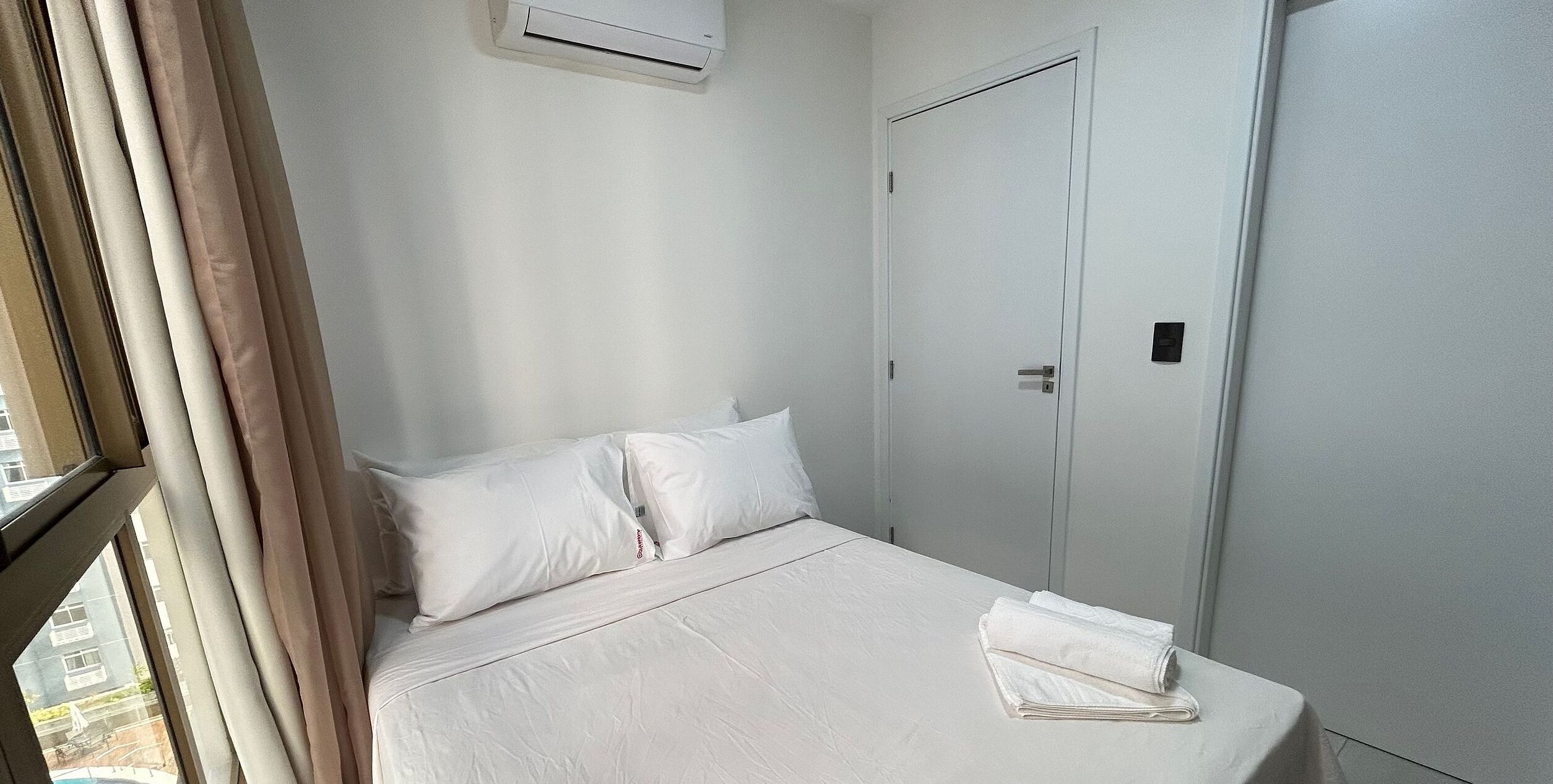 Une chambre d’hôtel avec un lit, des rideaux blancs, une porte fermée et un appareil de climatisation.