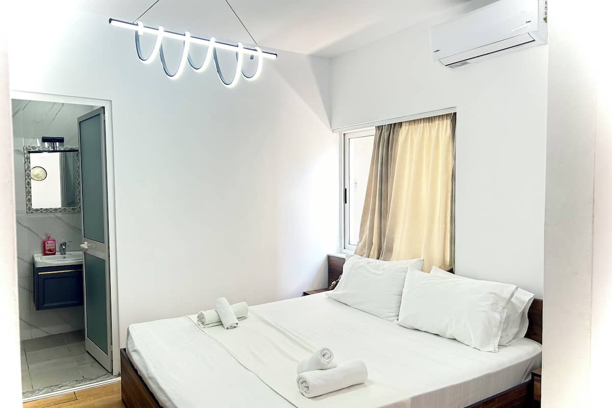 Habitación de hotel con cama, mesita de noche, espejo, ventana con cortinas y equipo de aire acondicionado.