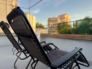 Ein schwarzer, klappbarer Stuhl mit Mesh-Rückenlehne und Armlehnen, platziert auf einem Balkon mit Blick auf Gebäude und Bäume.