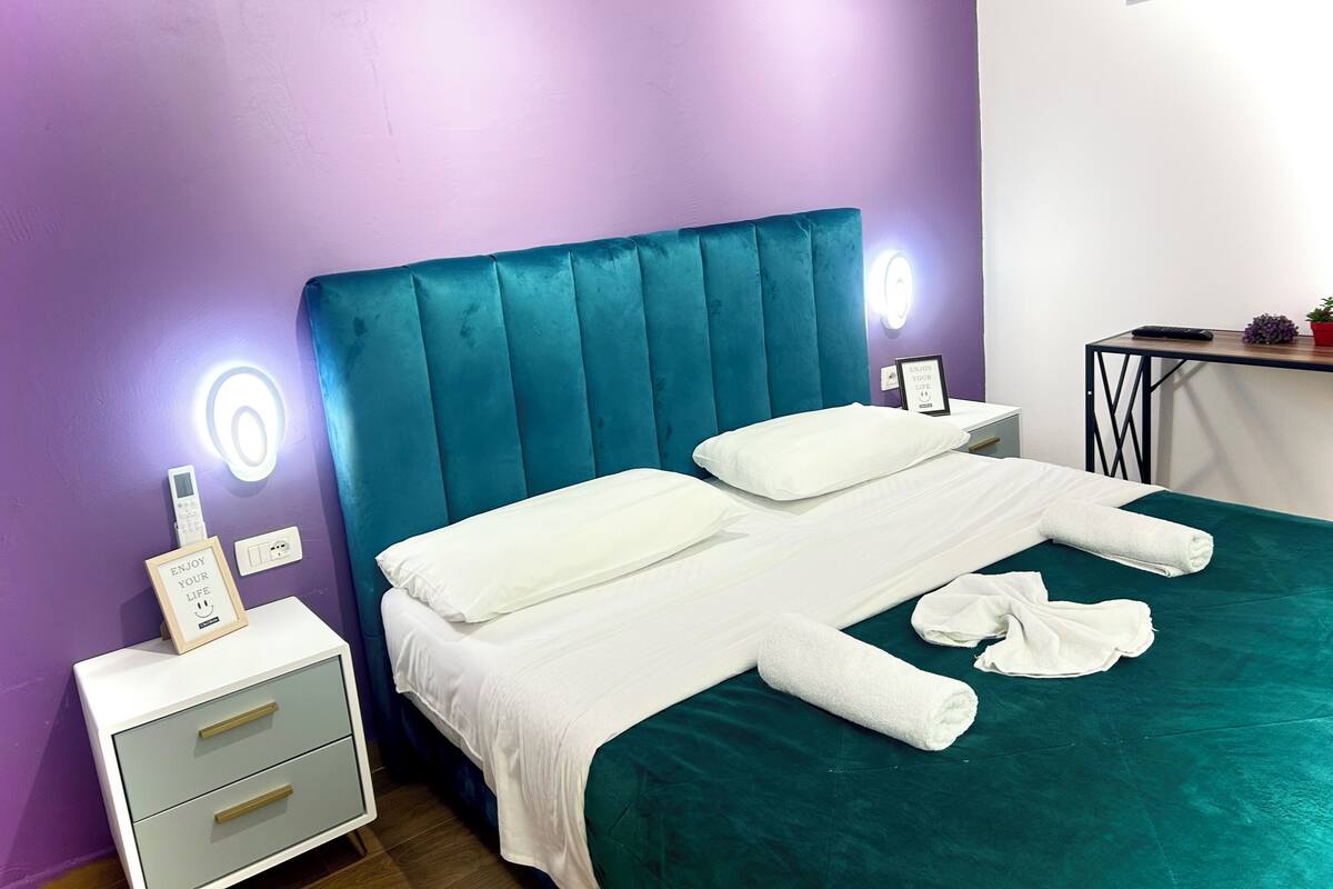 Una cama bien hecha con sábanas blancas y un cabecero color turquesa en una habitación con paredes moradas.