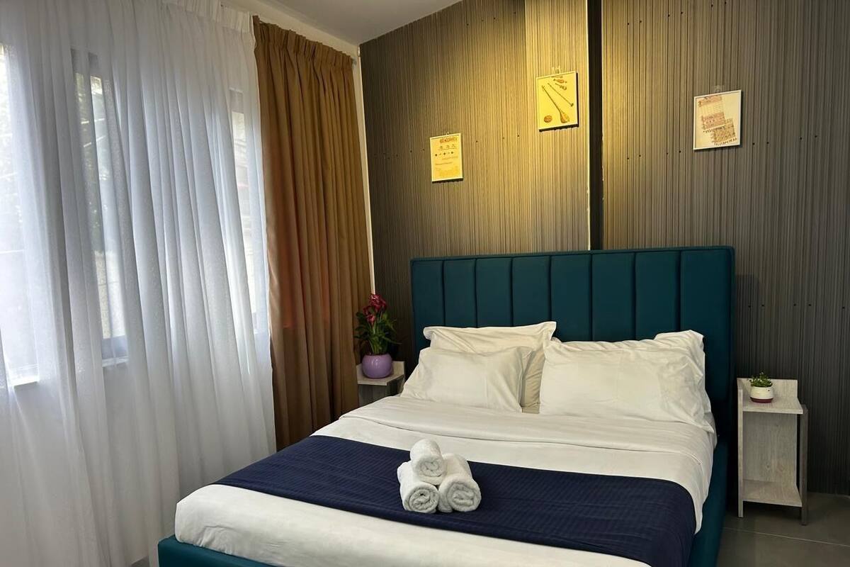 Habitación de hotel con cama, cortinas, mesita de noche, planta en maceta y letreros en la pared.