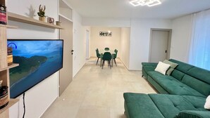 A modern living room with a green sofa, a flat-screen TV mounted on the wall, a dining area with a table and chairs, and a hallway leading to a door. - Rosoglio Poreč 300m from sea, new apartment 2024 (Poreč)