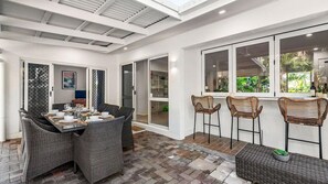 A dining area with a table set for four, surrounded by wicker chairs, and a view of greenery through a window.