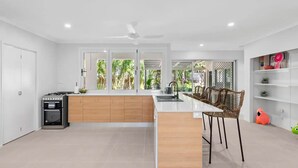 A modern kitchen with a central island, a dining area with wicker chairs, and a view of greenery through large windows. - Belle Escapes Sandpipers Beach House (Palm Cove)