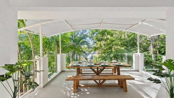 An outdoor dining area with a wooden table and benches, surrounded by white walls and glass panels, overlooking a lush garden.