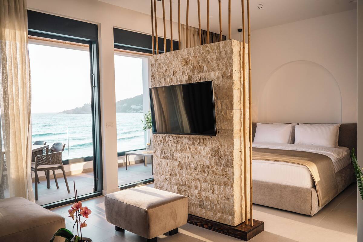 A modern bedroom with a stone accent wall, a flat-screen TV, a bed, and a view of the ocean.