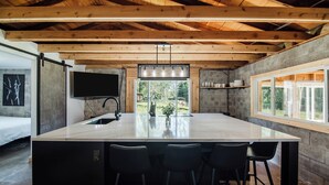 A modern kitchen with a large island, black bar stools, a built-in sink, and a window overlooking a garden. - The Cottage (Bellaire)