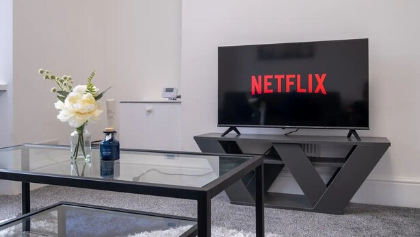 Un salon moderne doté d’un grand téléviseur à écran plat affichant le logo de Netflix, d’une table basse en verre et d’un vase rempli de fleurs.