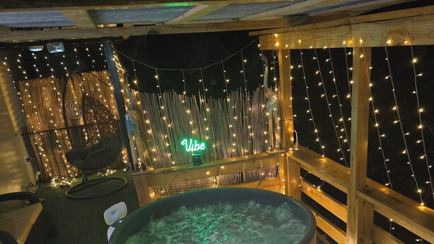 A hot tub surrounded by wooden railings and string lights, with a neon sign reading "Vibe" in the background.