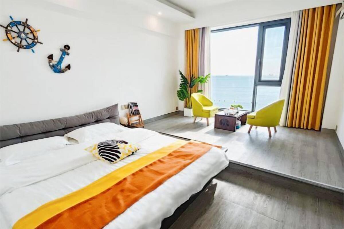 A modern hotel room with a large bed, a view of the sea, a yellow chair, and a small table with a plant.