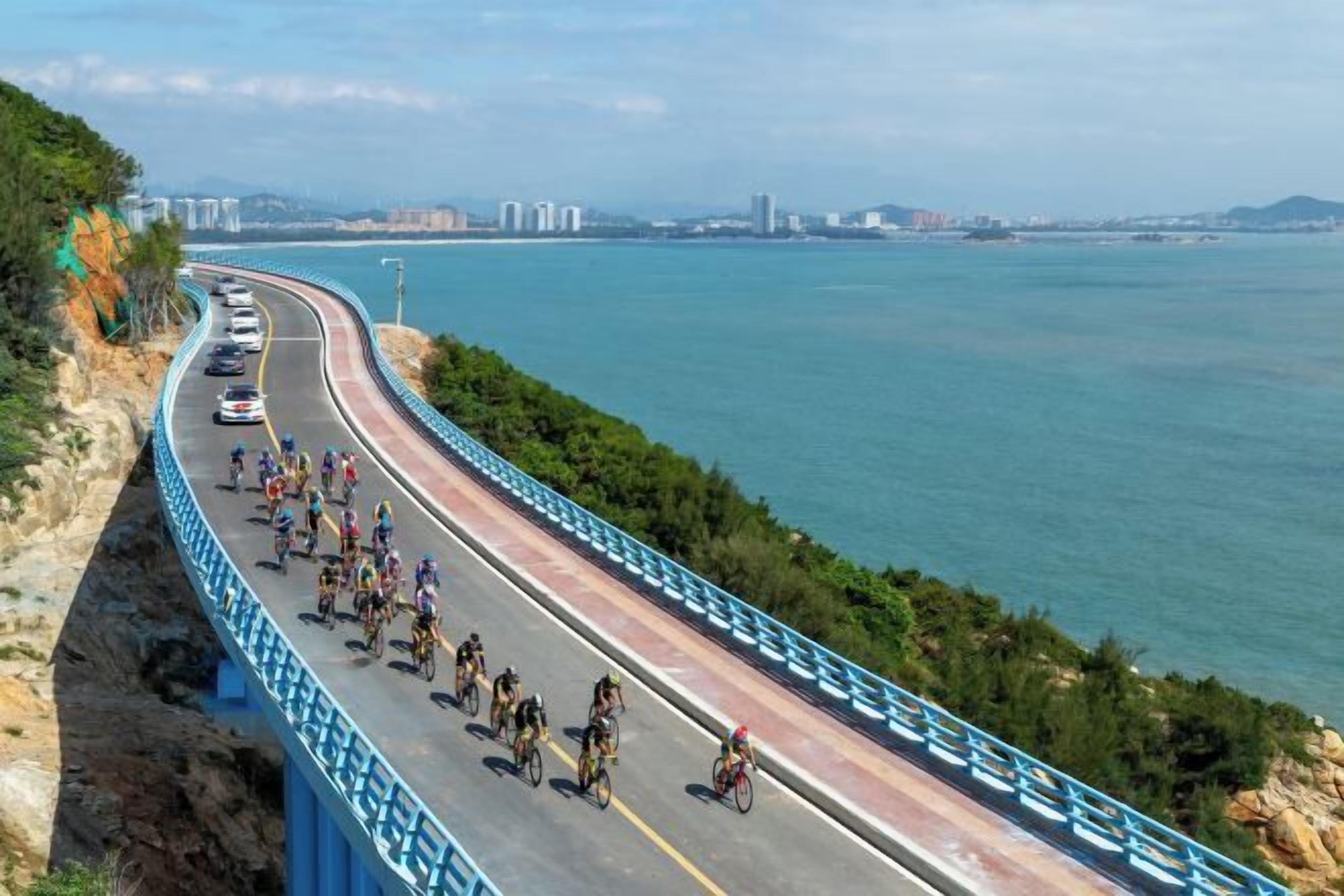 Une route côtière, un groupe de cyclistes, un pont et, au loin, la silhouette d’une ville.