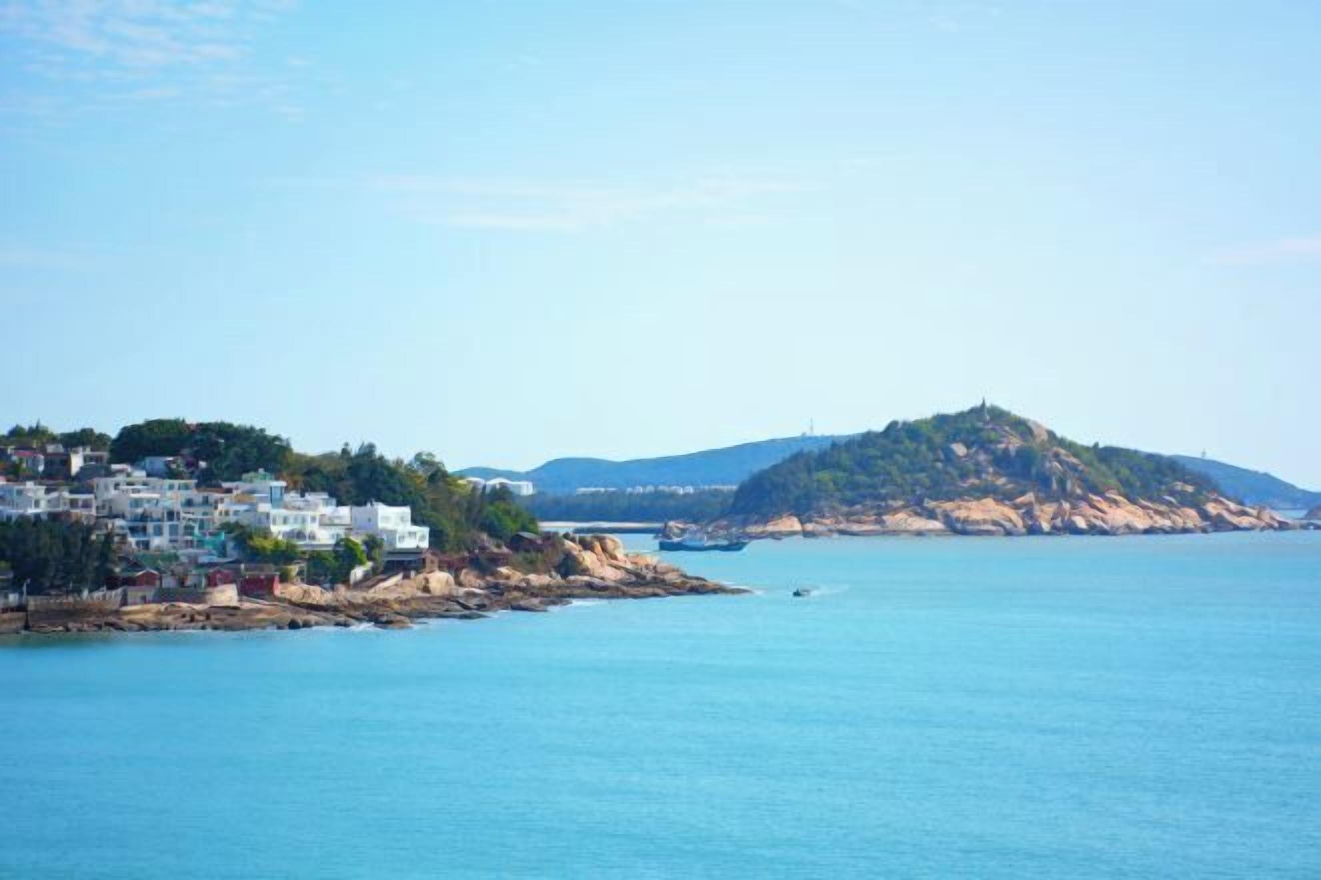 A coastal town with buildings along the shore, a prominent hill, and calm sea.