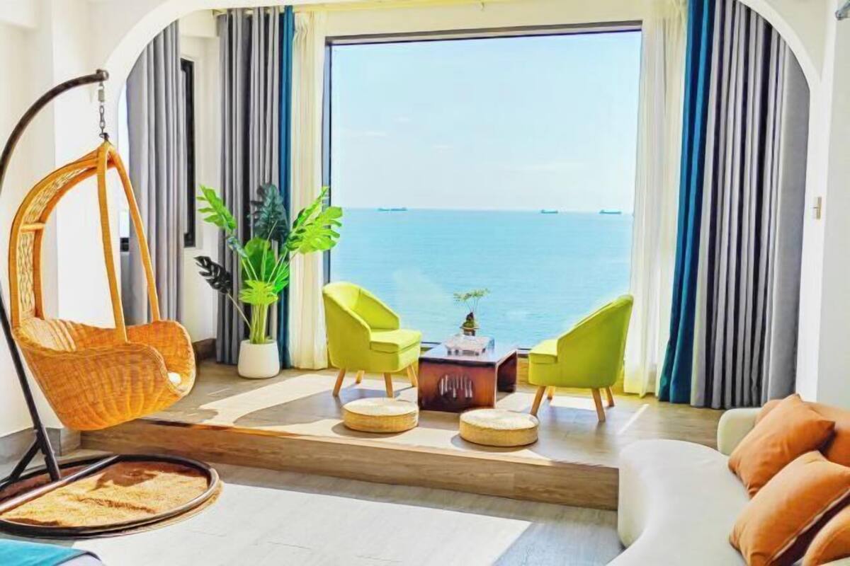 A room with a view of the sea, a hanging chair, two green chairs, a small table, and a sofa.