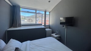 A hotel room with a bed, a window with a view of a cityscape, a flat-screen TV mounted on the wall, and a small bedside table with a lamp. - M & B Backpackers (Cape Town)