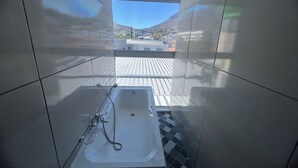 A modern bathroom with a bathtub, a shower, and a window offering a view of a cityscape and mountains. - M & B Backpackers (Cape Town)