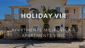 Een gebouw met twee verdiepingen, voorzien van balkons en een stenen gevel, met de tekst 'HOLIDAY VIR APARTMENTS MILJKOVICA & APARTMENTS VIRIC'.