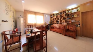 A spacious living room with a wooden dining table and chairs, a sofa set, and a bookshelf filled with various items.