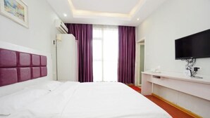 A hotel room with a large bed, a flat-screen TV mounted on the wall, and a window with purple curtains.
