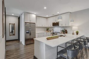 A modern kitchen with white cabinets, a stainless steel refrigerator, a central island with bar stools, and pendant lighting.