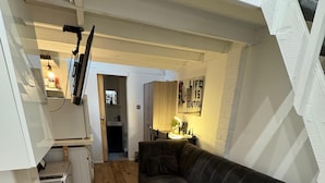 Ein kompakter Wohnbereich mit Holzboden, einem grauen Sofa, einer kleinen Küchenzeile mit Kühlschrank und Mikrowelle sowie einer Tür zu einem weiteren Raum.