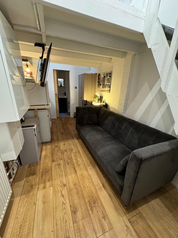 Un espace de vie compact doté d’un plancher en bois, d’un canapé gris, d’un petit coin cuisine avec un réfrigérateur et un four à micro-ondes, et d’une porte donnant sur une autre pièce.