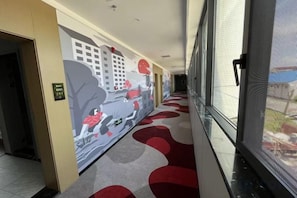 A hotel corridor with a red carpet, a mural on the wall, and a door on the left.