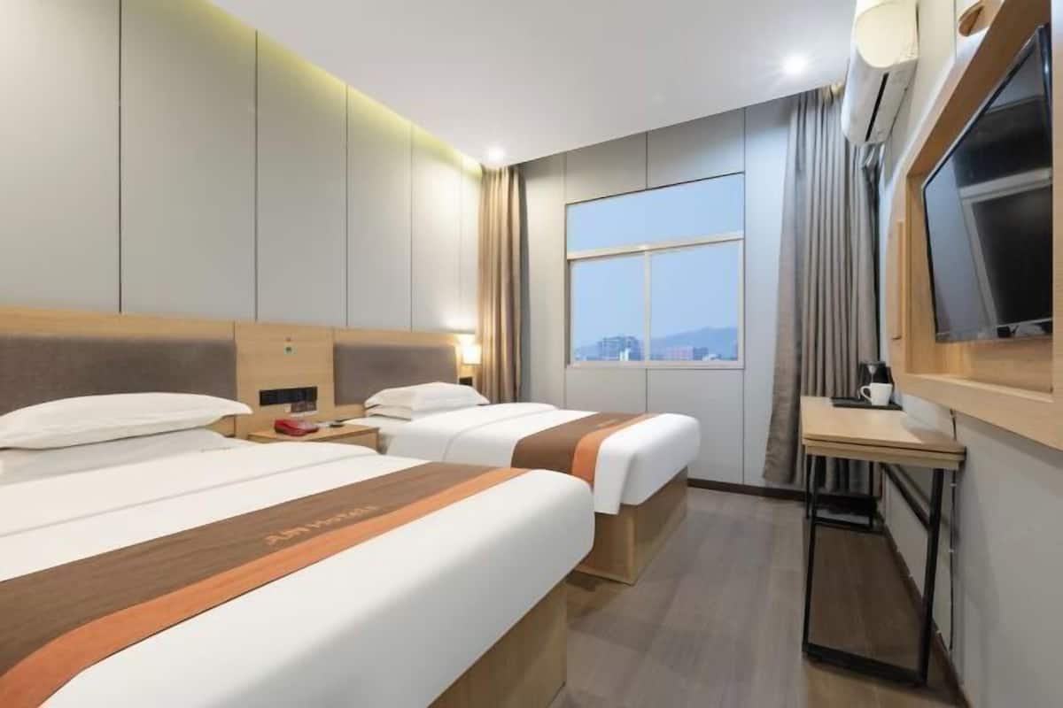 A hotel room with two beds, a desk, a TV, and a window with a city view.