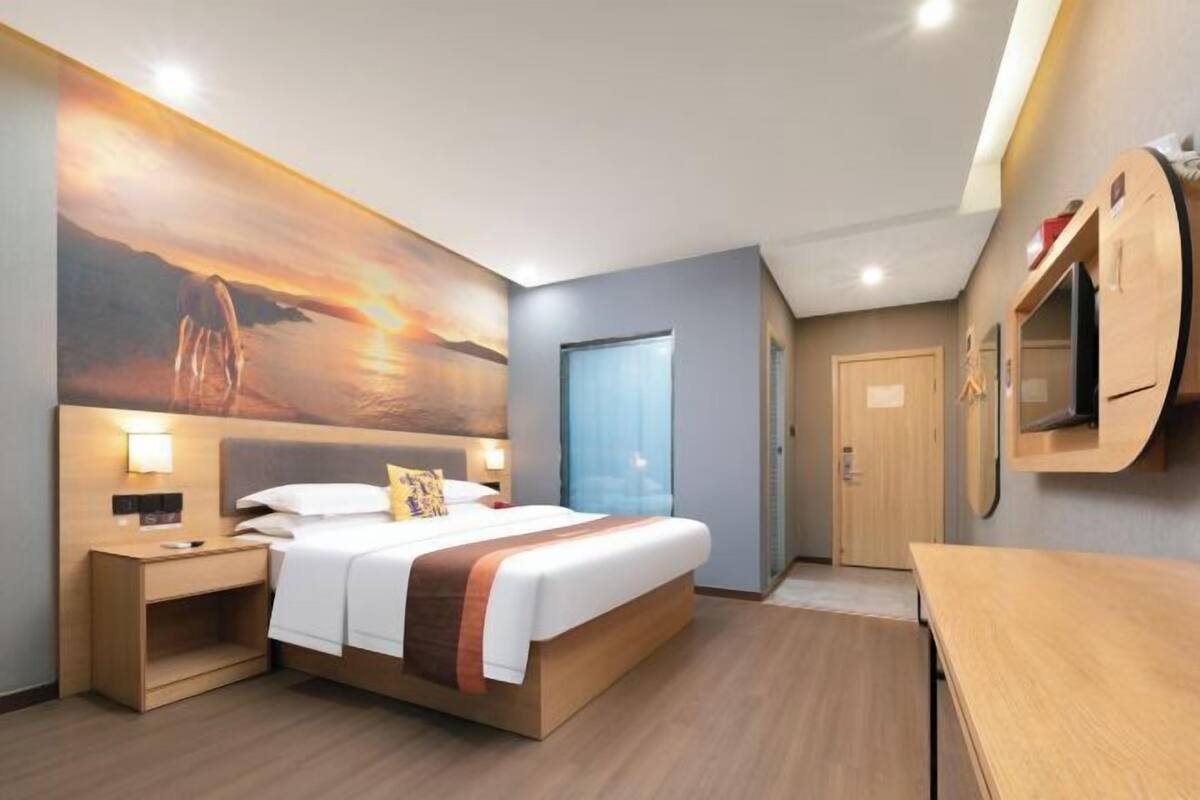 A modern hotel room with a large bed, bedside tables, a TV, and a painting of a sunset.
