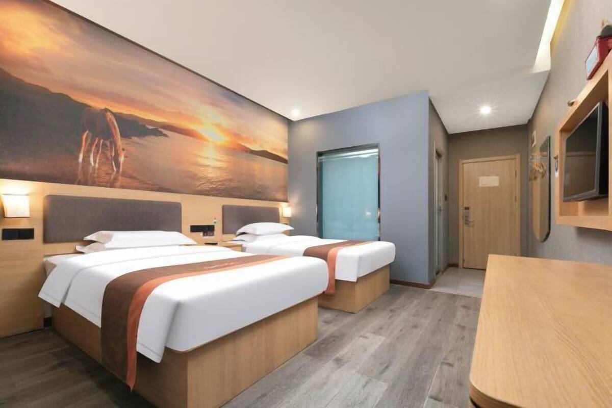 A hotel room with two beds, a large painting of a sunset, a wooden table, and a TV mounted on the wall.