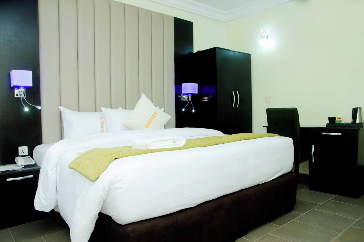 A hotel room with a large bed, a nightstand with a phone, a desk with a lamp, and a wardrobe.