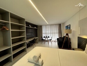 Ein modernes Hotelzimmer mit einem großen Bett, einem an der Wand montierten Fernseher, einem Schreibtisch mit Stühlen, einem Esstisch mit Stühlen und einem Schrank mit Regalen.